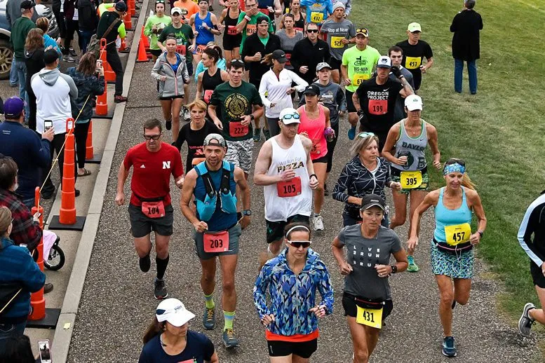 A group of runners in various athletic wear are running forward on a paved path, with spectators visible on the left side of the image.