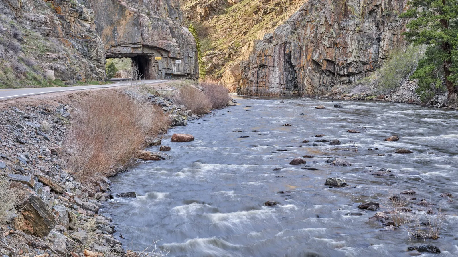 A scenic view of a road passing through a tunnel next to a flowing river with rocky cliffs.