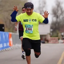 A male runner in a neon yellow shirt and black shorts, with his arms raised in celebration, crosses the finish line of a road race.