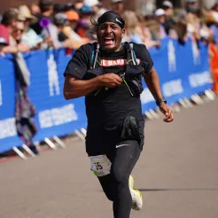 A male runner with a bandana and determined expression runs towards the camera, wearing a black t-shirt and compression leggings, with spectators blurred in the background.