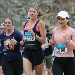 Three female runners in the foreground, wearing race bibs and running on a road course.
