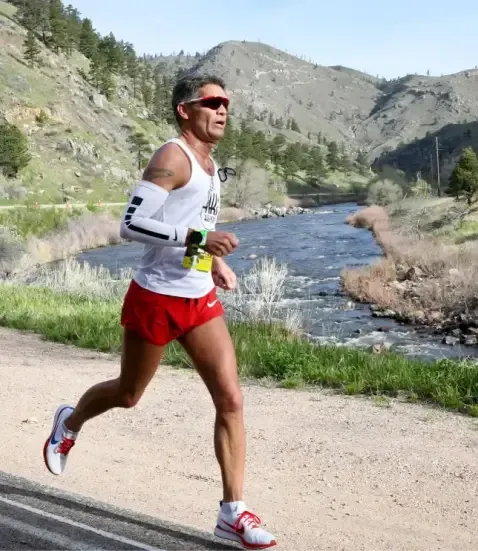 A male runner in red shorts and a white tank top runs along a path next to a river with mountains in the background.