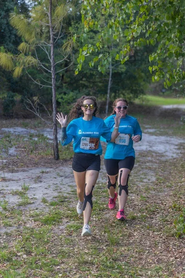 Two women running in a race, wearing blue shirts and bib numbers, smiling and waving.