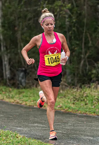 A female runner with blonde hair in a ponytail, wearing a pink tank top and black shorts, runs on a paved path with trees in the background. She is wearing a bib with the number 1045.
