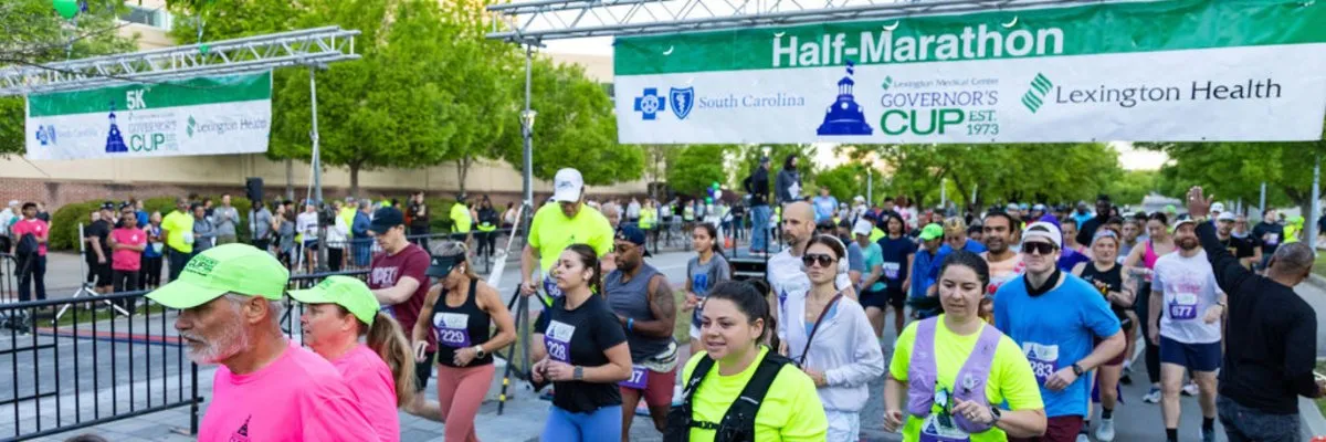 A large group of runners are gathered at the start line of the Governor's Cup Half-Marathon, with race banners overhead.