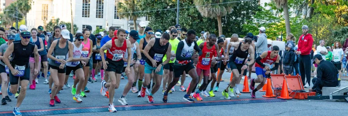 A group of runners are lined up at the start of a road race, with some in motion as the race begins.