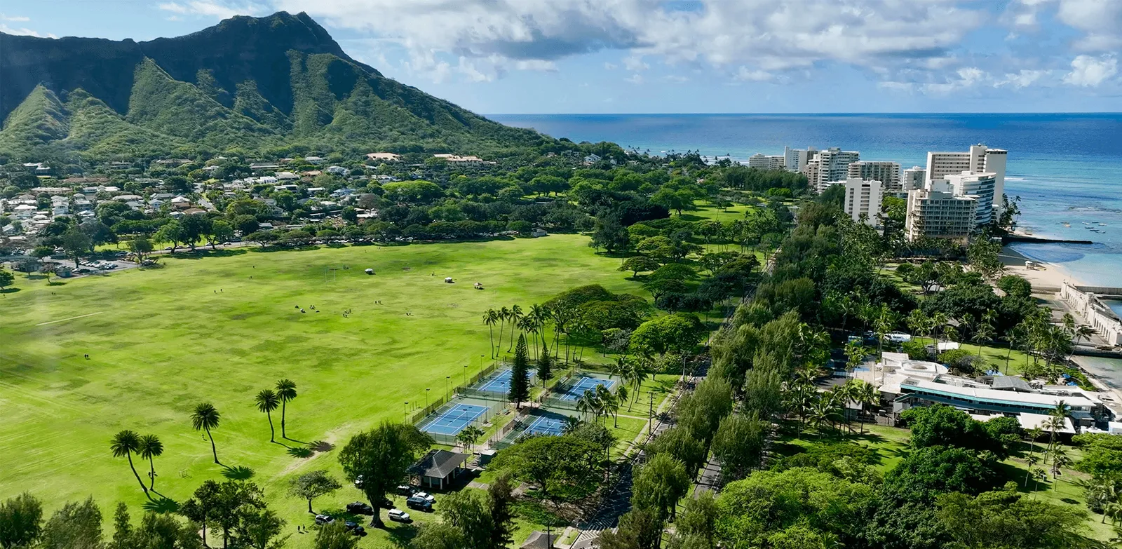 Aerial view of the Hapalua Hawaii Half Marathon course with Diamond Head mountain, lush green fields, palm trees, urban buildings, and the ocean.