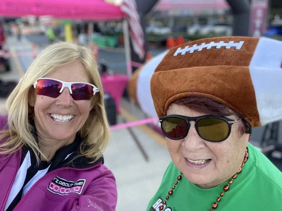 Two smiling women at a race, one wearing a football hat and the other wearing sunglasses.