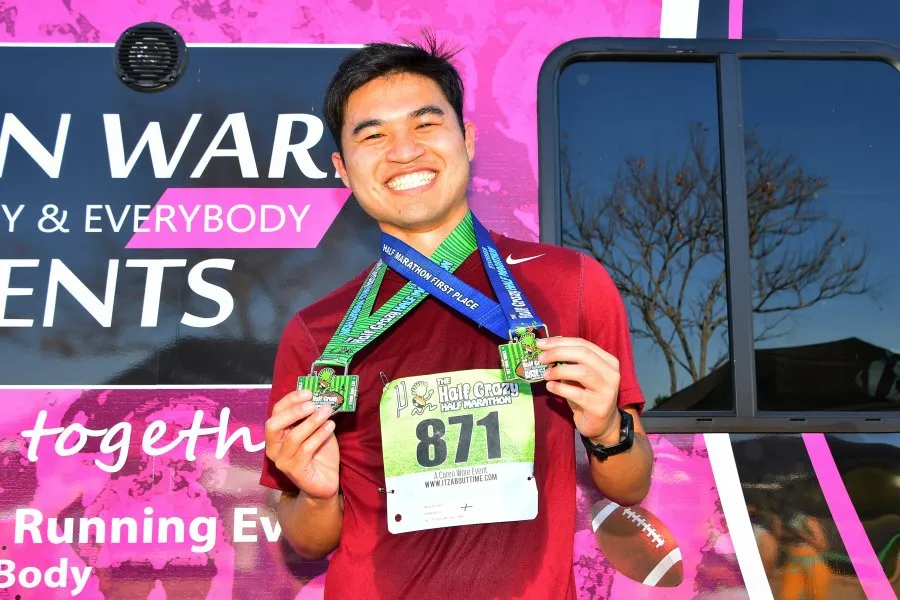 A smiling runner holds up two medals, one blue and one green, in front of a pink and black banner.