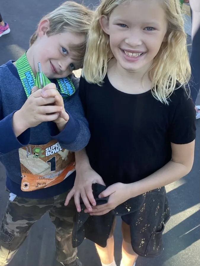Two smiling children, one wearing a race medal and bib, posing for a photo after a race.