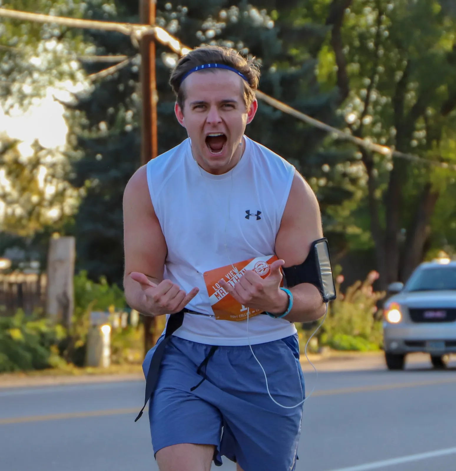 A male runner with an excited expression, mouth open wide, running on a road with trees and a car in the background. He is wearing a white tank top with a race bib and blue shorts.