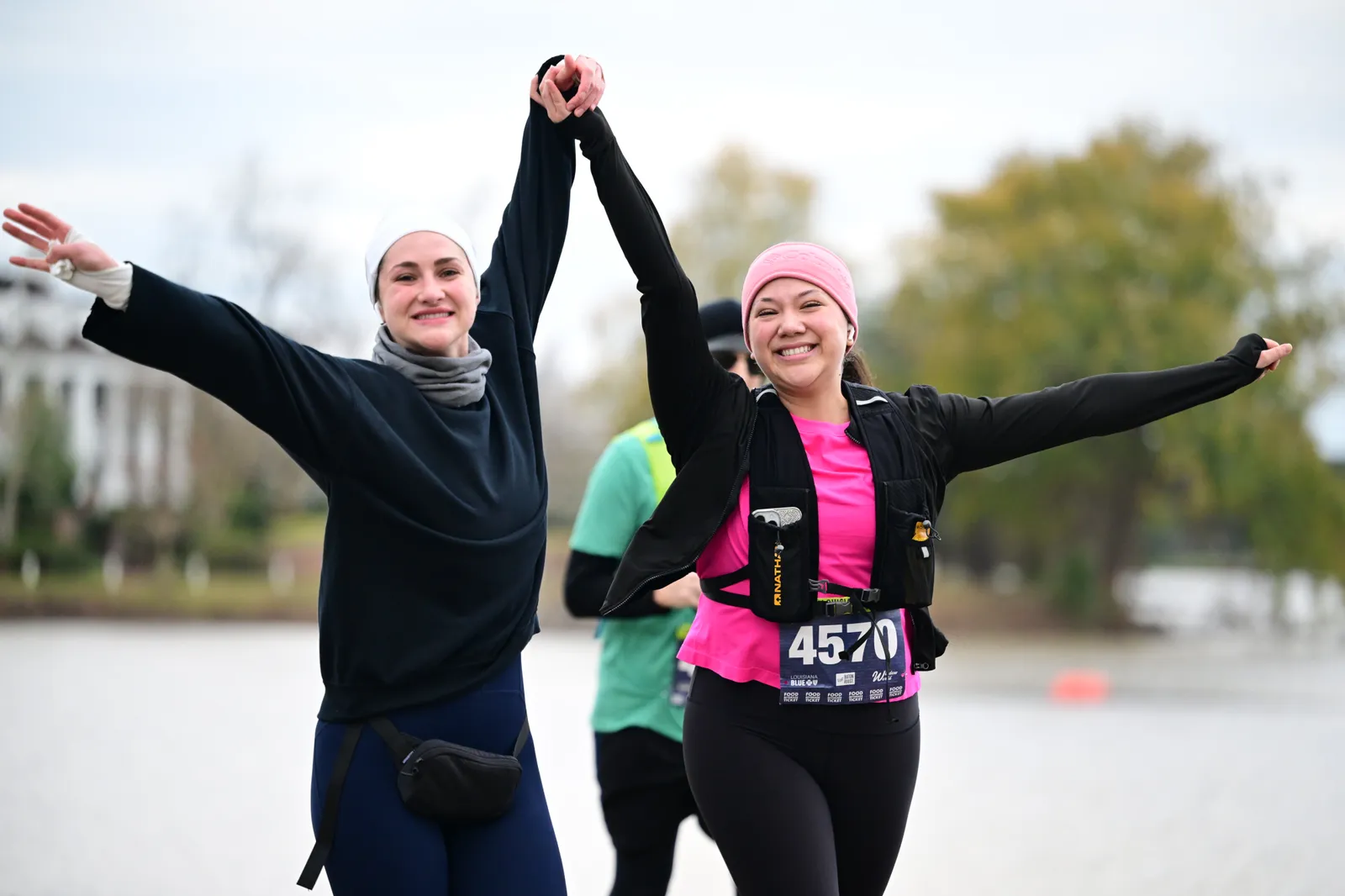 Two women with their arms raised in celebration, holding hands, wearing race bibs.