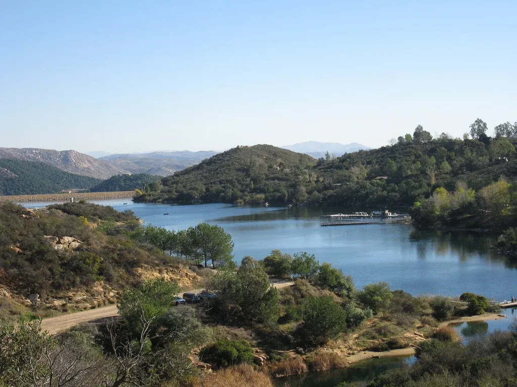 Scenic view of a lake with rolling hills and trees, a road is visible in the foreground.