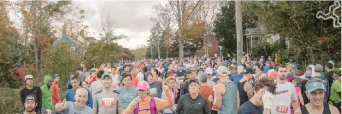 A large crowd of runners gathered before a race, with trees and houses in the background.