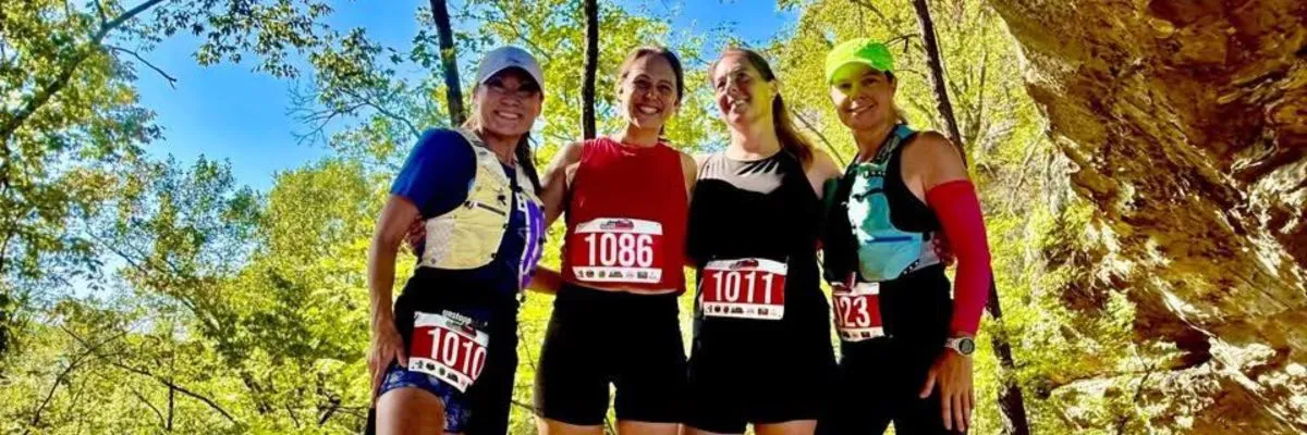Four women smiling and posing for a photo during a trail race, wearing bib numbers.