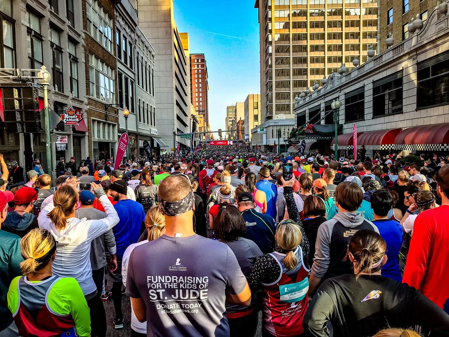 A large crowd of runners gathered on an urban street, looking towards the start line of a race.