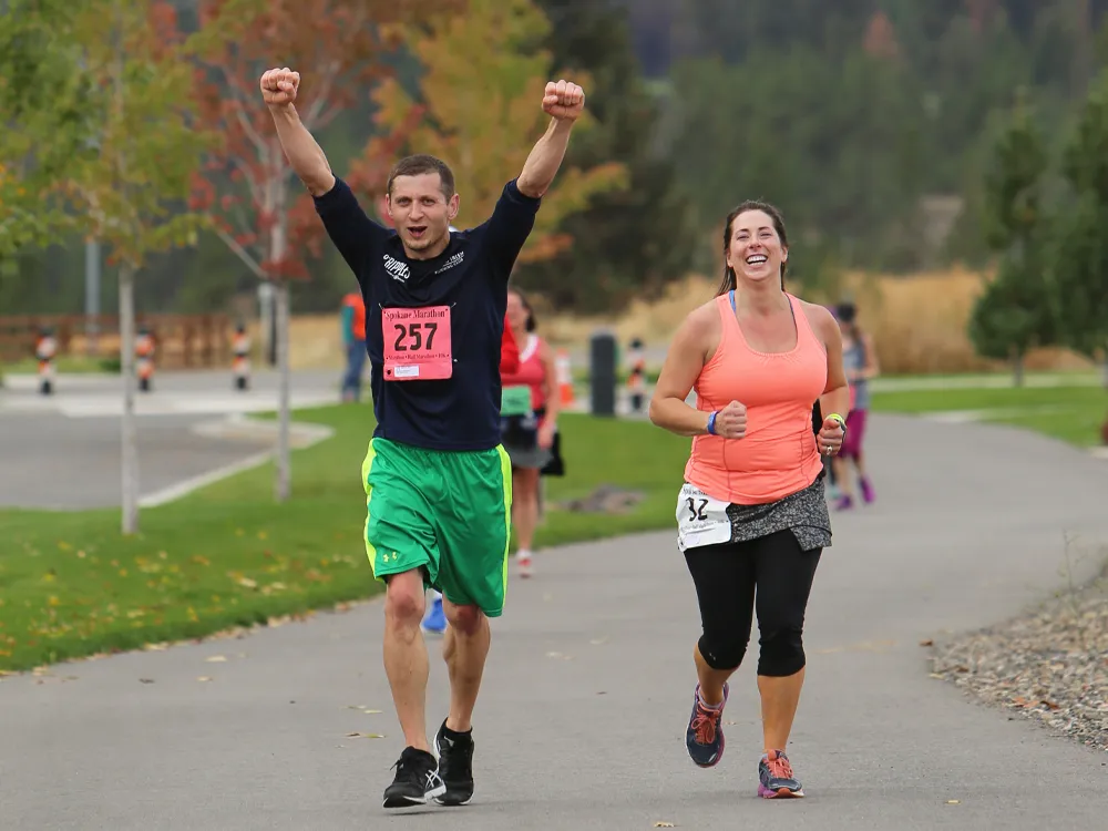 A male runner with his arms raised in celebration and a female runner smiling as they run towards the finish line.