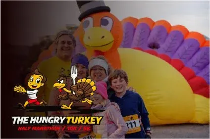 A group of people, including children, pose for a photo in front of a large inflatable turkey at The Hungry Turkey Half Marathon/10K/5K race.