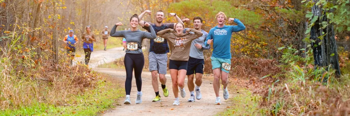 A group of runners smile and flex their muscles while running on a dirt path through an autumn forest.