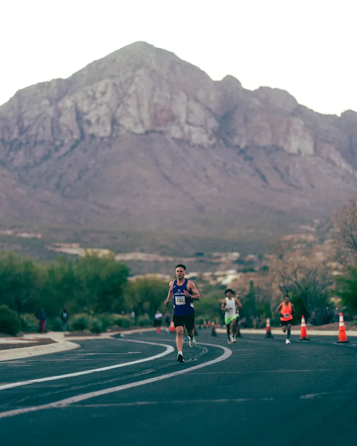 A male runner in a blue tank top and black shorts runs on a paved road with a large, rocky mountain in the background.