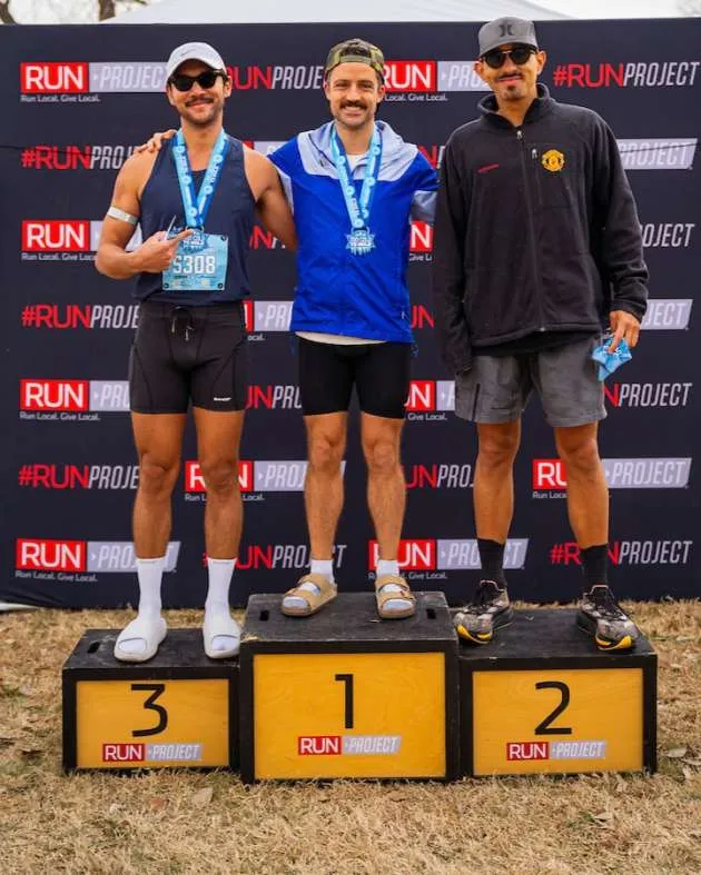 Three male runners stand on a podium, with the first, second, and third place winners holding medals.