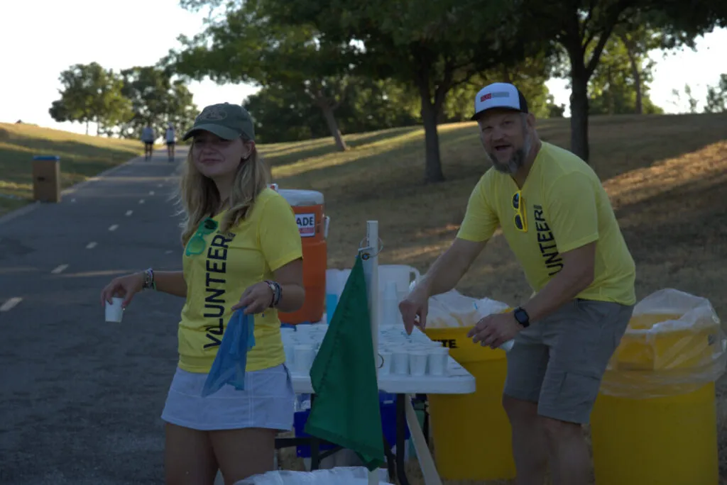 Two volunteers, a woman and a man, wearing yellow shirts that say "VOLUNTEER" are standing at a water station on a paved path during a race.