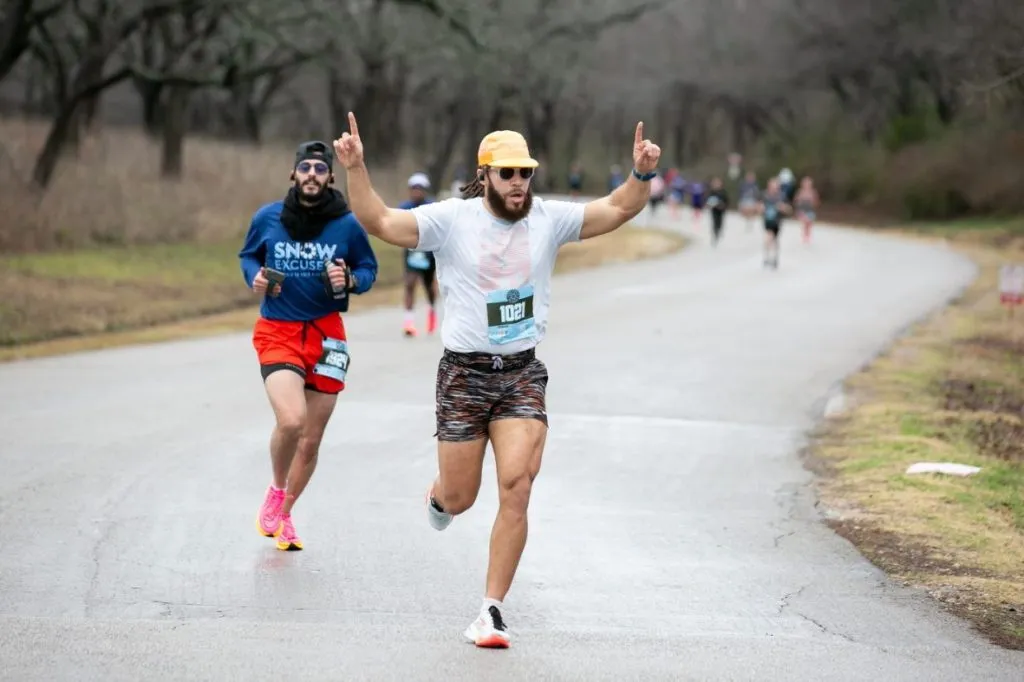 A runner celebrates with arms raised while running on a paved road, with other runners in the background.