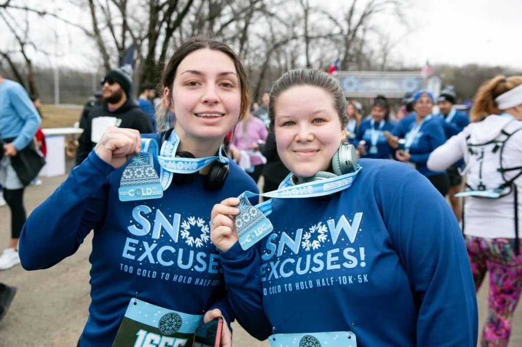Two smiling women wearing blue 'Snow Excuses' long-sleeve shirts and race medals, with other runners in the background.