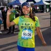 A female runner smiles and waves at the camera, wearing a yellow and green tie-dye race shirt with bib number 5605, a blue baseball cap, and blue leggings.