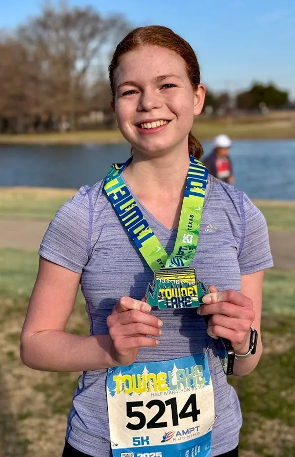 A young woman smiles proudly while holding her Towne Lake Half Marathon medal and wearing her race bib.