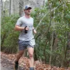 A male runner wearing a blue baseball cap and grey t-shirt runs on a wooded trail, with a bib number visible on his shorts.