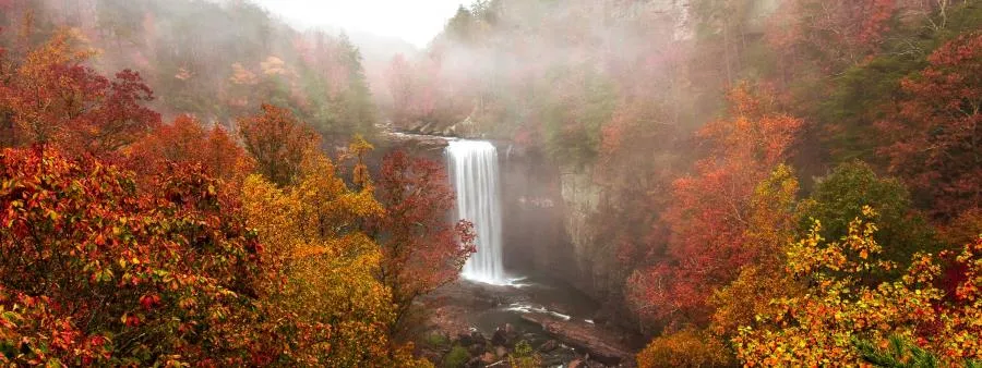 A waterfall cascades down a rocky cliff surrounded by trees displaying vibrant autumn colors of red, orange, and yellow, with mist rising in the background.