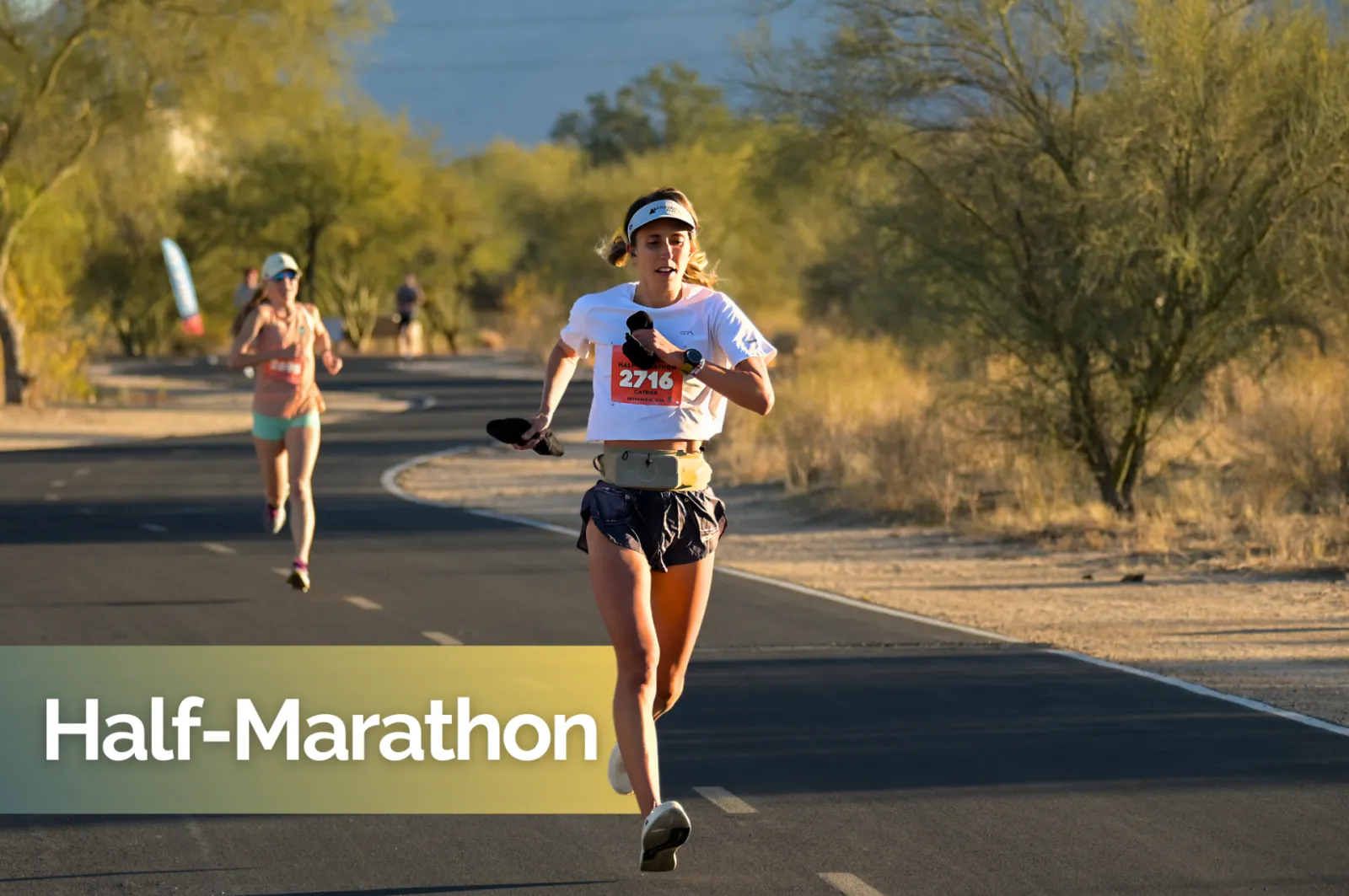 A female runner in the foreground, wearing bib number 2716, runs on a paved road during a half marathon. Another runner is visible in the background.