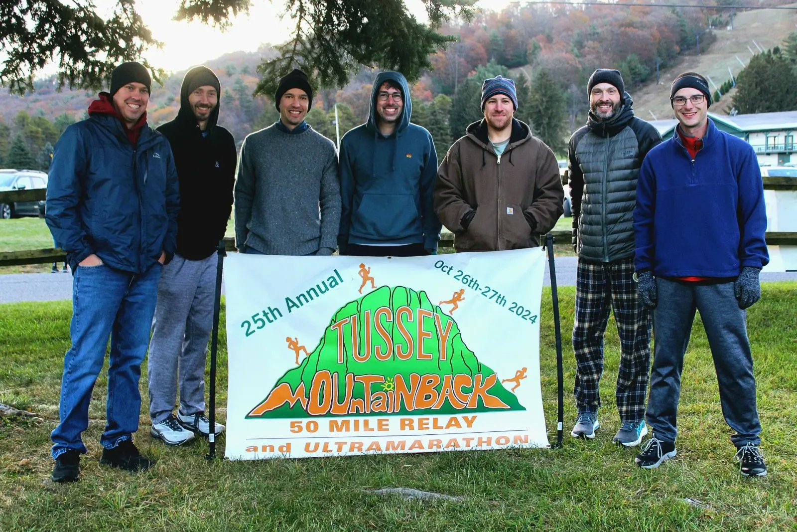 A group of seven men smile and pose in front of a banner for the Tussey Mountainback 50 Mile Relay and Ultramarathon.