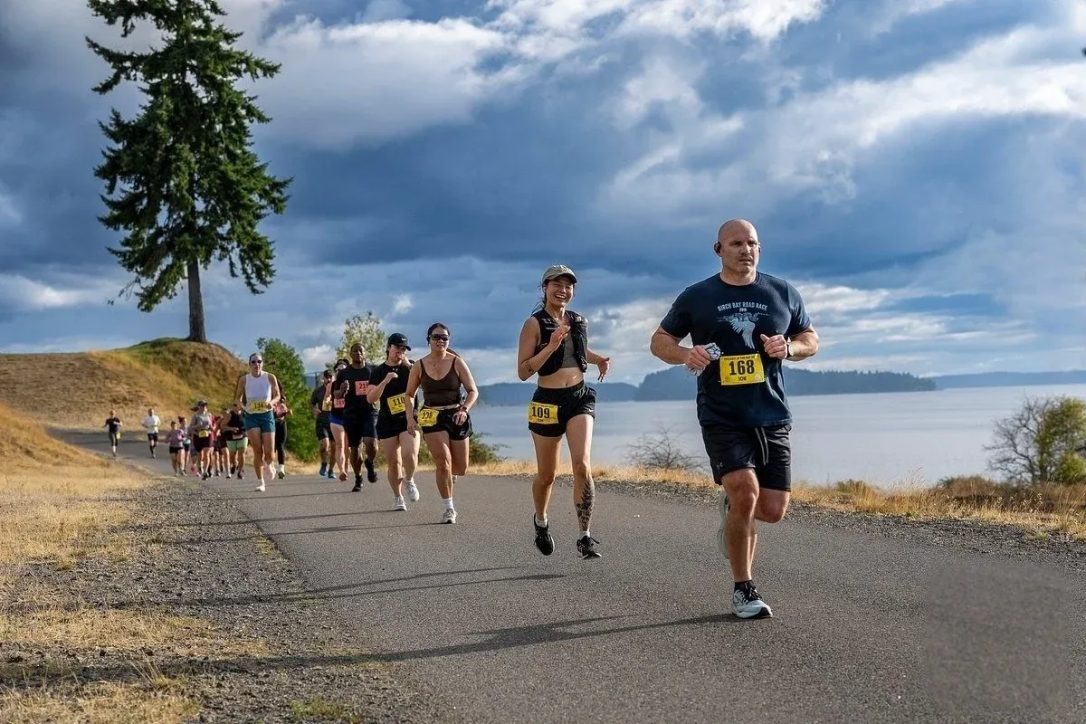 A group of runners on a paved road with a scenic waterfront view in the background.