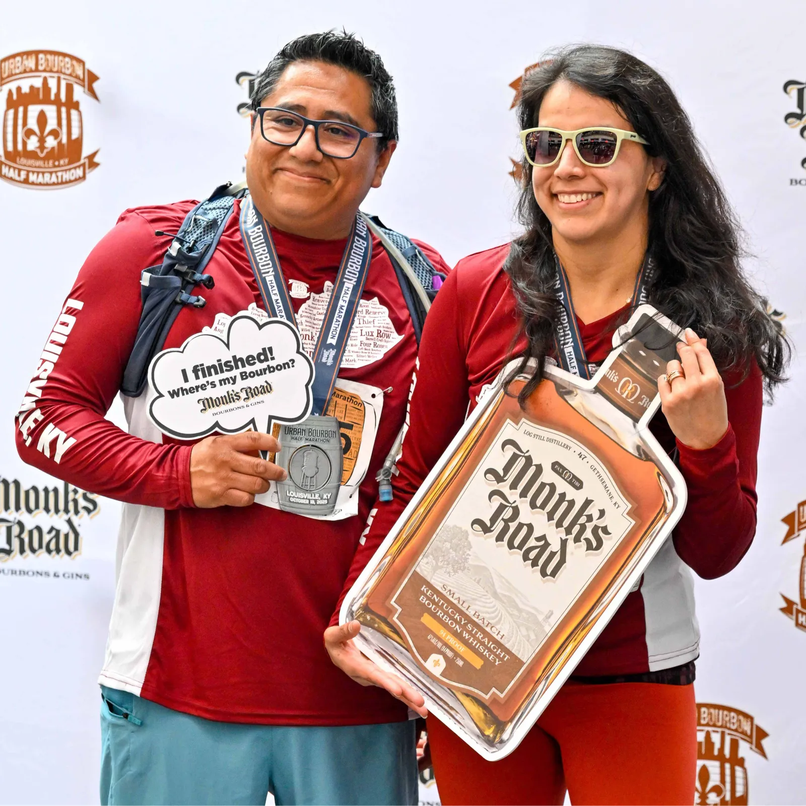 Two smiling runners, one holding a 'I finished! Where's my Bourbon?' sign and a medal, and the other holding a large cutout of a bourbon bottle, pose for a photo after completing the Urban Bourbon Half Marathon.