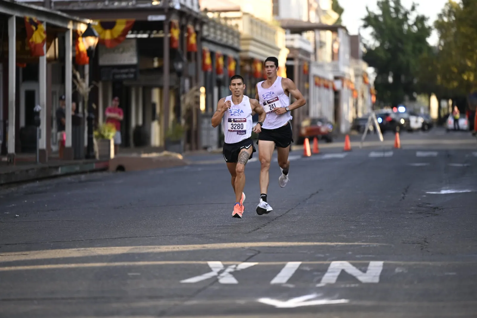 Two male runners race down a street lined with buildings and decorations, with police cars visible in the background.