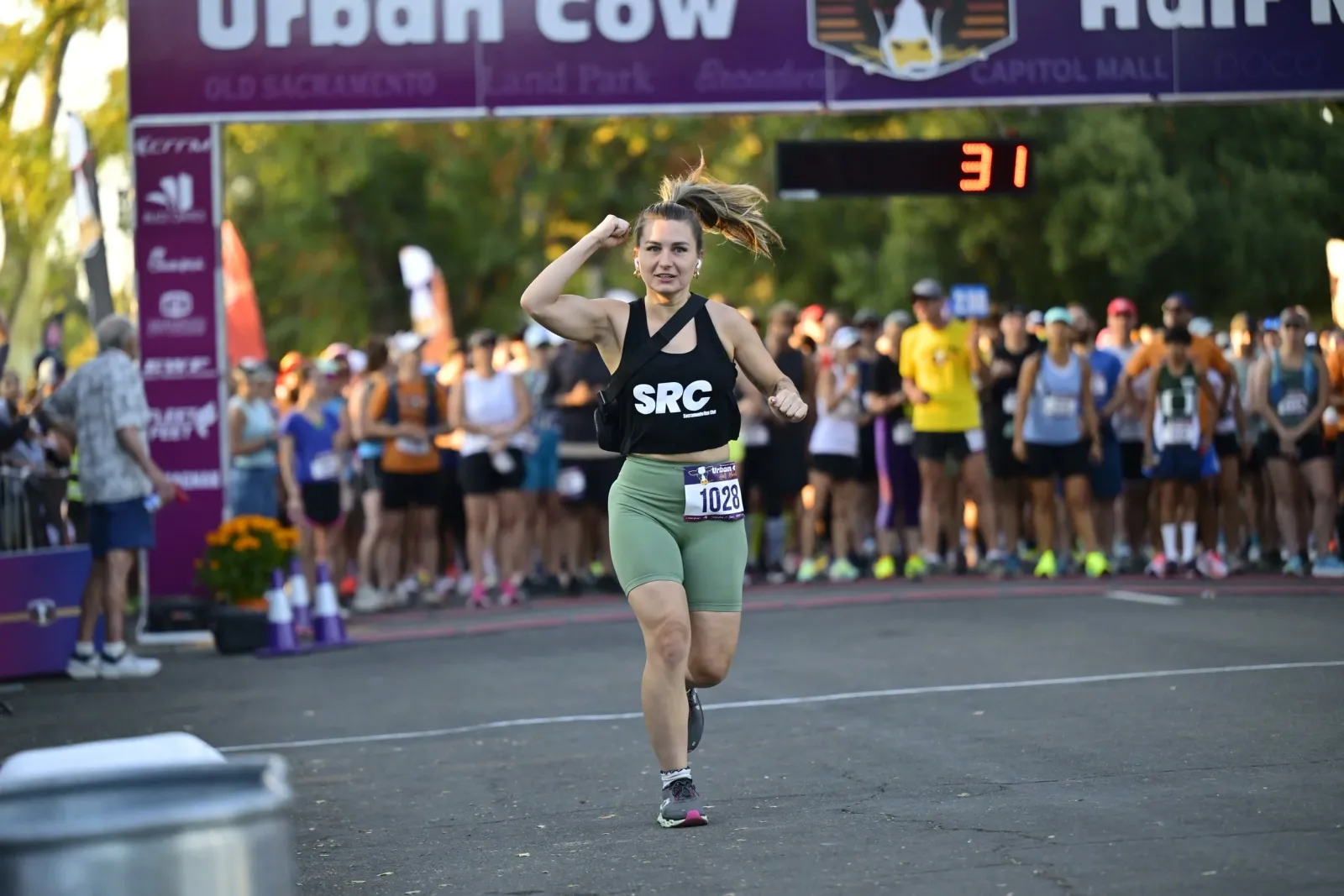 A female runner with her fist raised in celebration runs towards the start line of the Urban Cow Half Marathon, with other runners and race banners in the background.