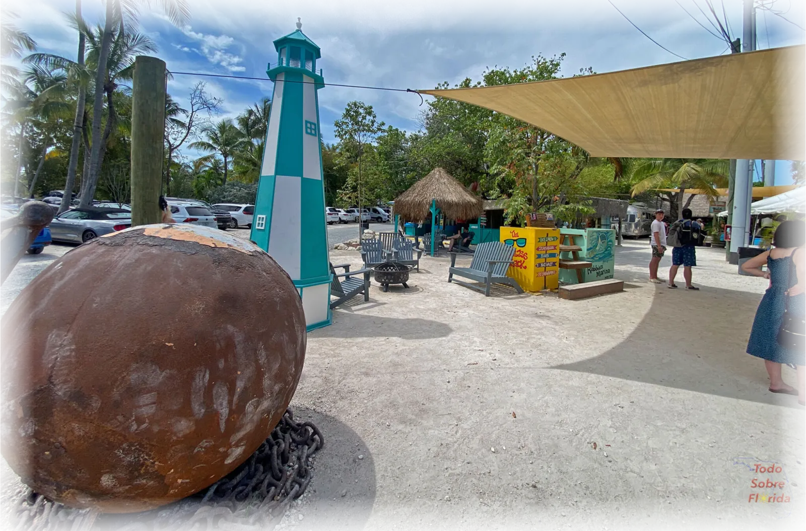 A scenic view of a waterfront area with a teal and white lighthouse, a large rusty anchor, palm trees, and people relaxing in chairs.