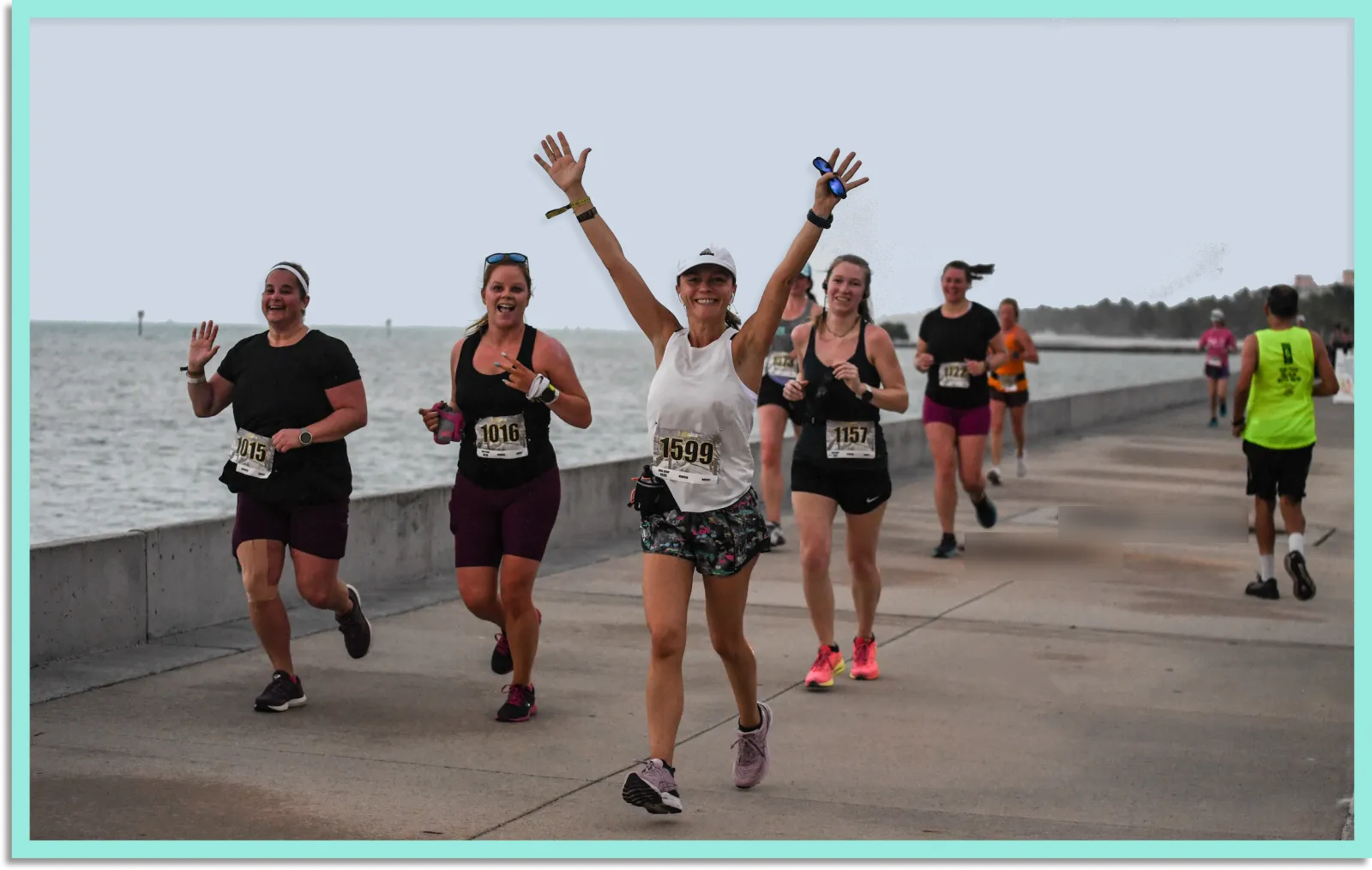 A group of women running a half marathon along a waterfront path, with one runner in the foreground raising her arms in celebration.