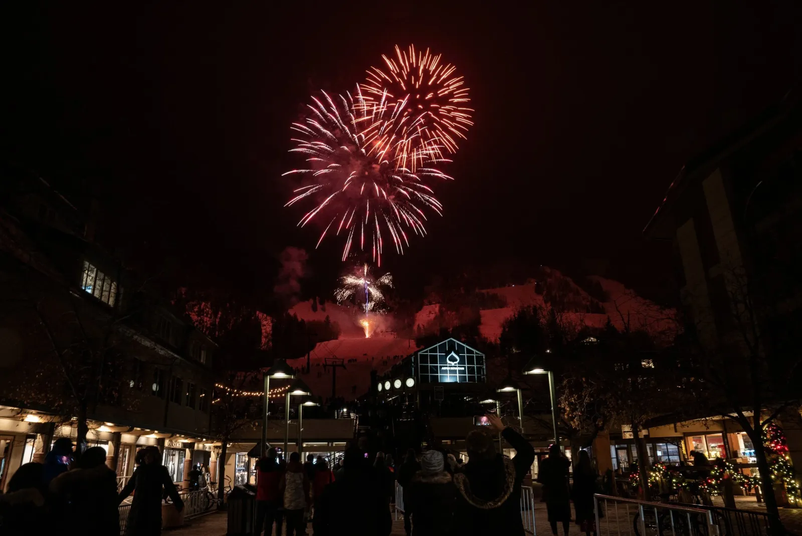 Fireworks explode in the night sky over a snowy mountain resort town, with a crowd of people watching from below.