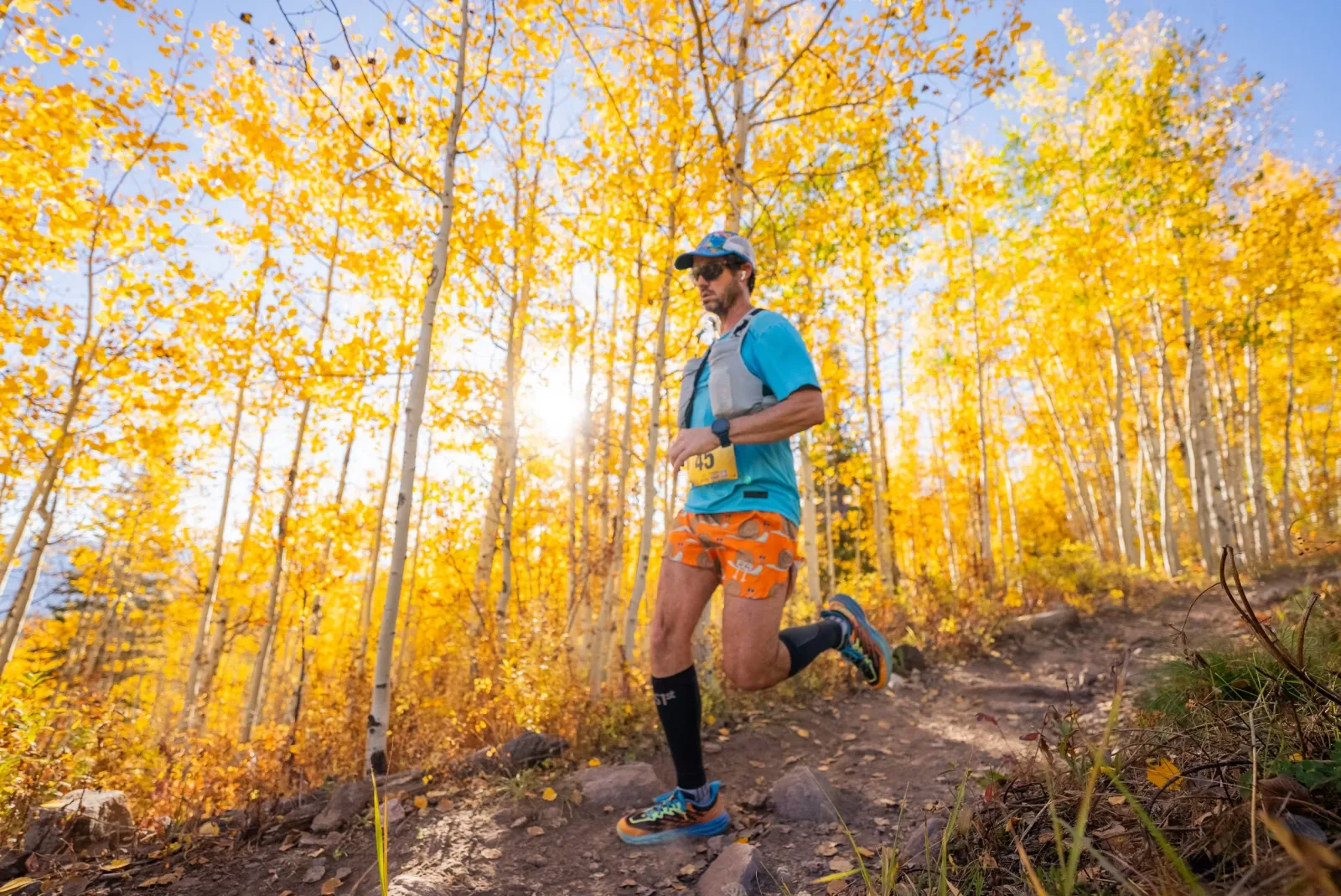 A male runner with a bib number 45 runs on a dirt trail through a forest with bright yellow aspen trees in the fall.