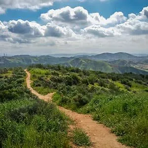 A winding dirt trail through green, rolling hills under a partly cloudy sky.