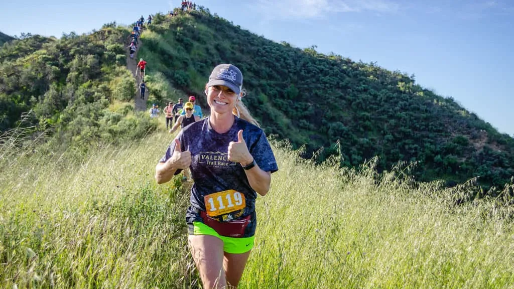 A female runner smiles and gives two thumbs up while running on a grassy trail, with other runners ascending a hill behind her.