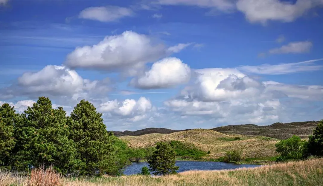 Scenic view of rolling sand hills with a small lake and trees under a cloudy blue sky.