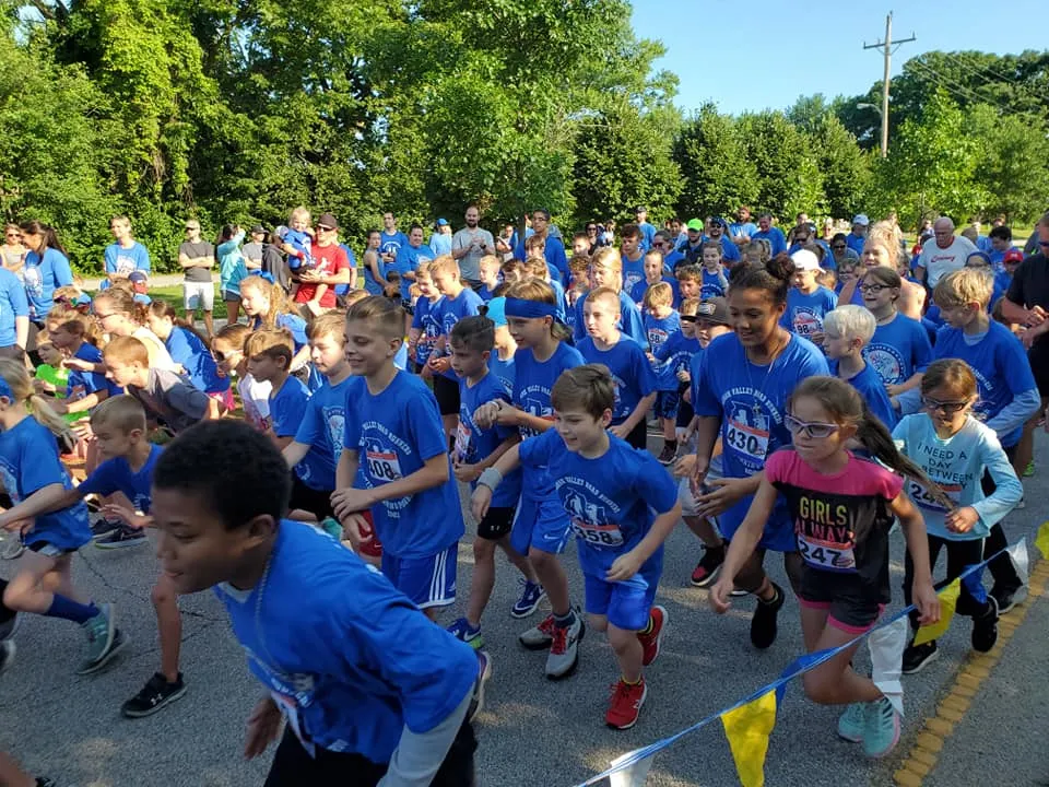 A large group of runners, mostly children, wearing blue shirts are lined up at the start of a race, with some beginning to run forward.