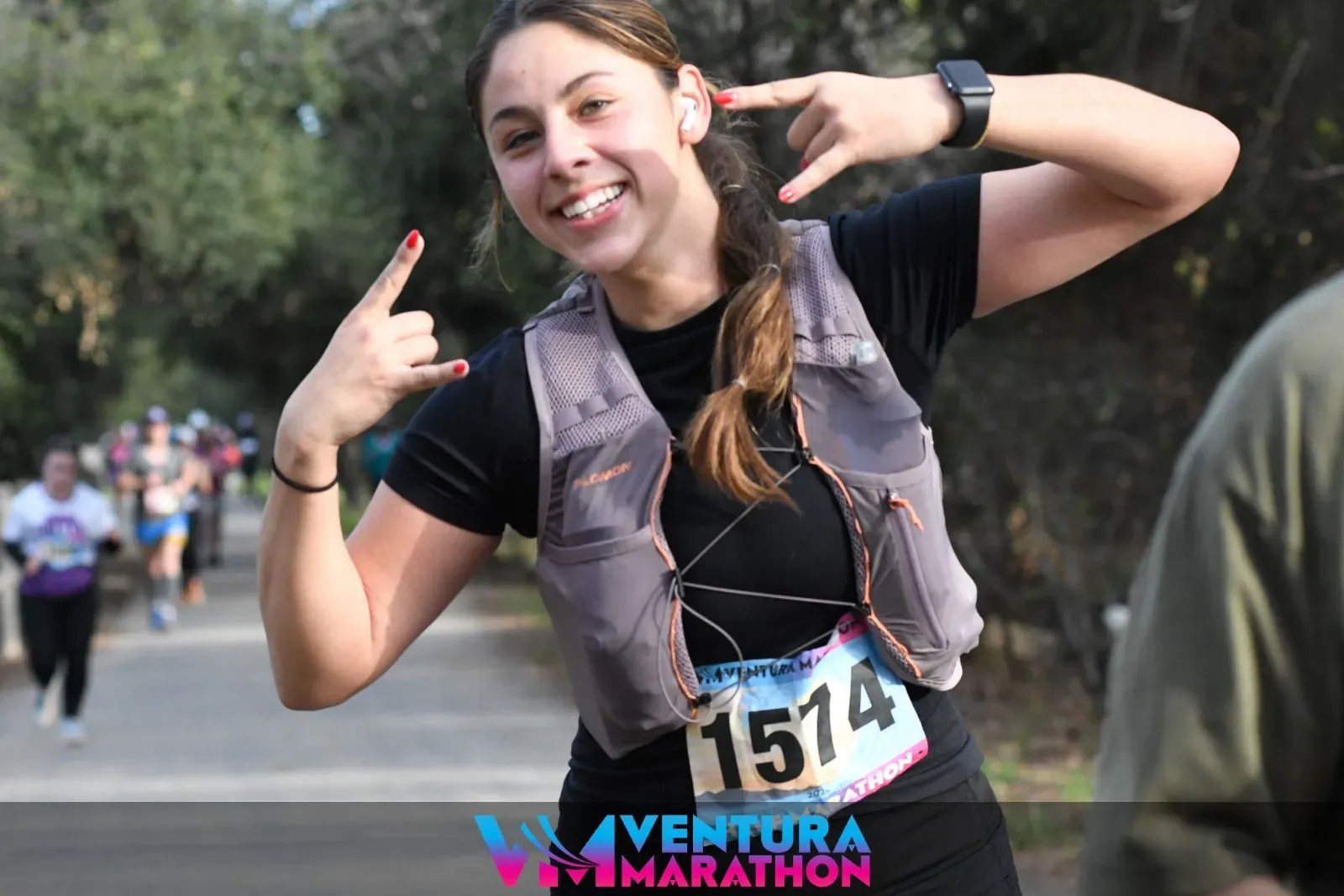 A smiling female runner with bib number 1574 makes a rock-on hand gesture while running.