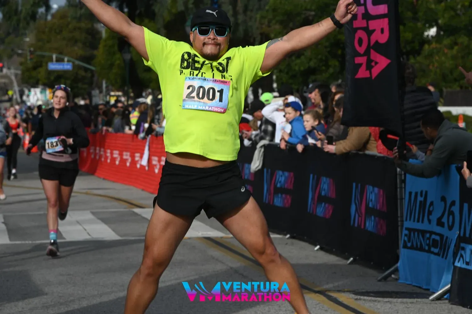 A runner in a bright yellow shirt with the number 2001 celebrates with arms outstretched at the finish line of the Ventura Marathon.