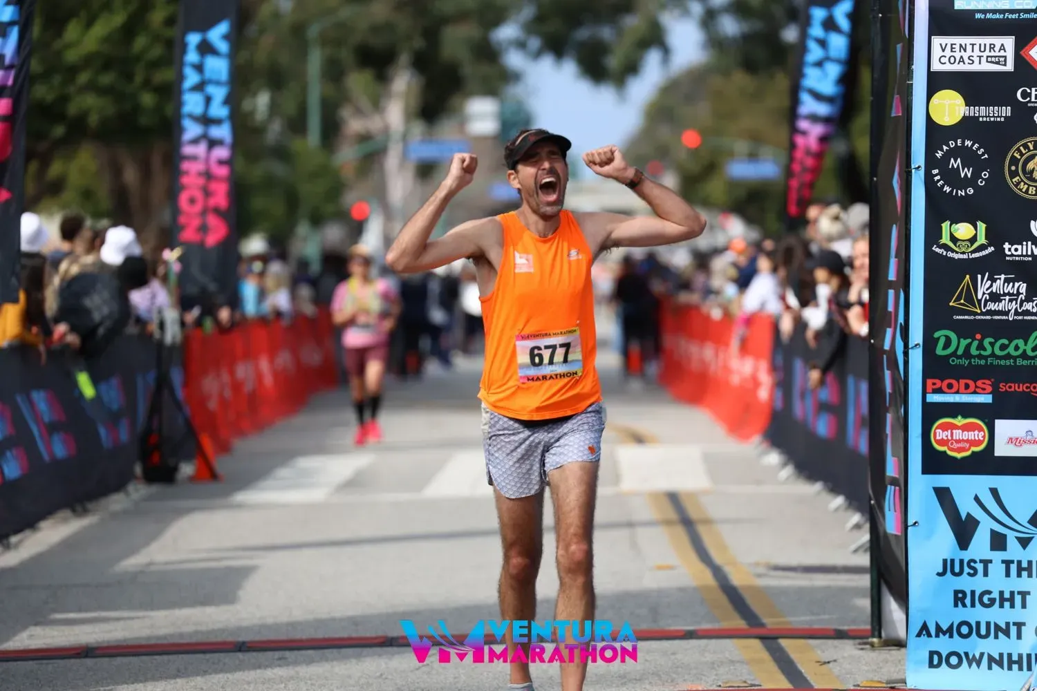 A male runner in an orange tank top and patterned shorts crosses the finish line of the Ventura Marathon with his arms raised in celebration.