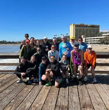 A group of runners posing for a photo on a wooden pier, with the ocean and a hotel in the background.